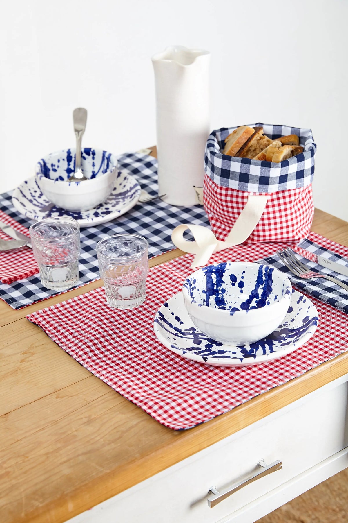 Two red and white gingham fabric napkins on a wooden table, set with plates, bowls, and a bread basket.