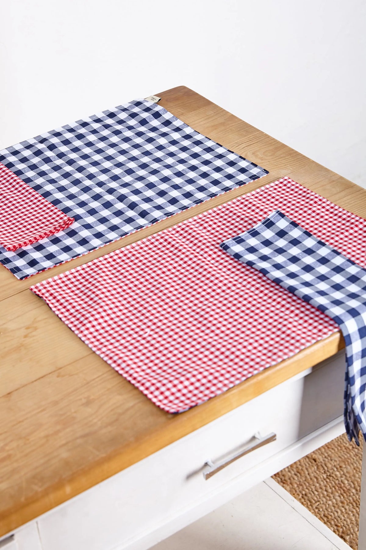 Pack de dos servilletas de tela con estampado de cuadros vichy en rojo y azul sobre mesa de madera clara.
