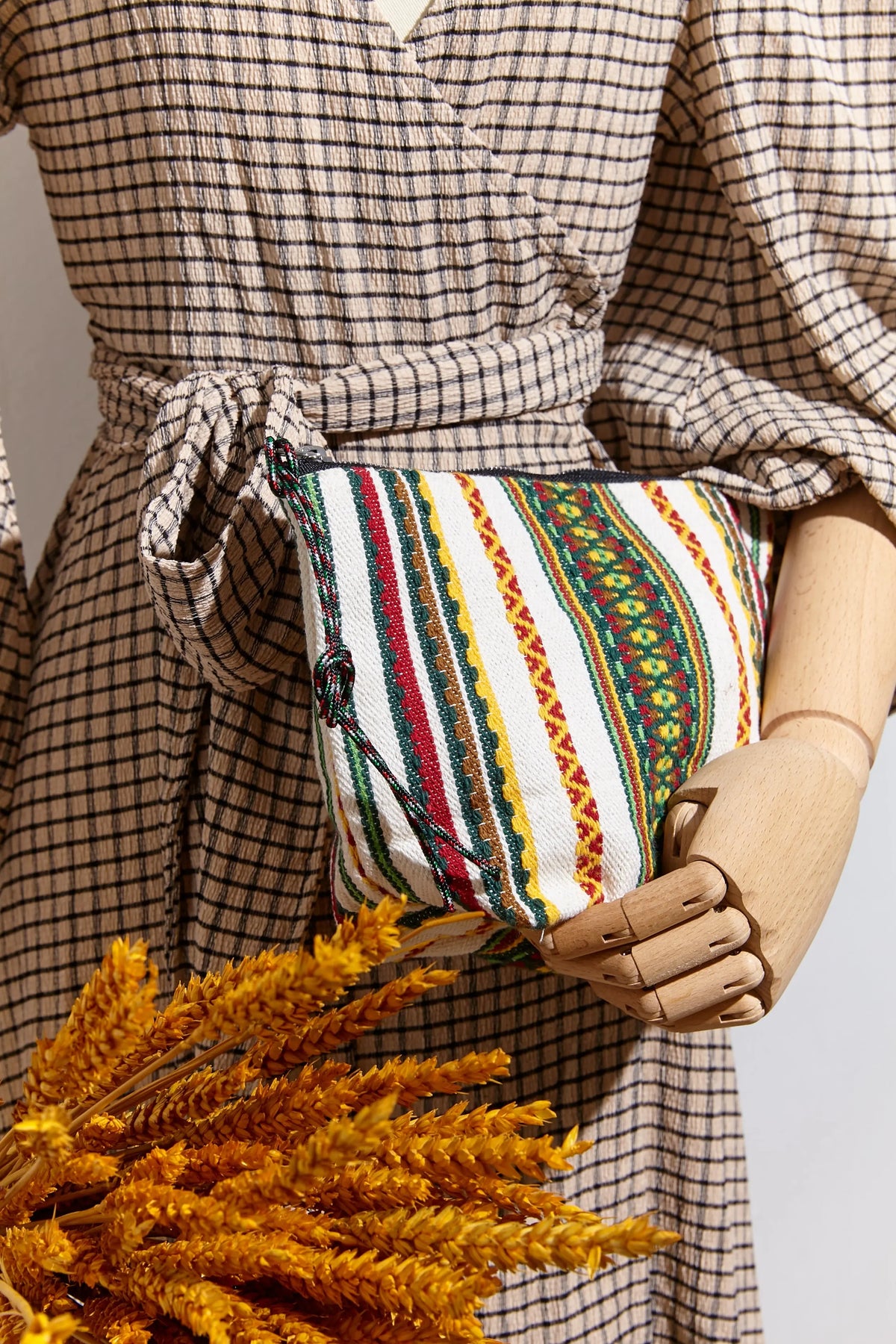 Neceser de tela cruda con rayas coloridas tejidas, sostenido por un maniquí. Diseño artesanal y cierre de cremallera.