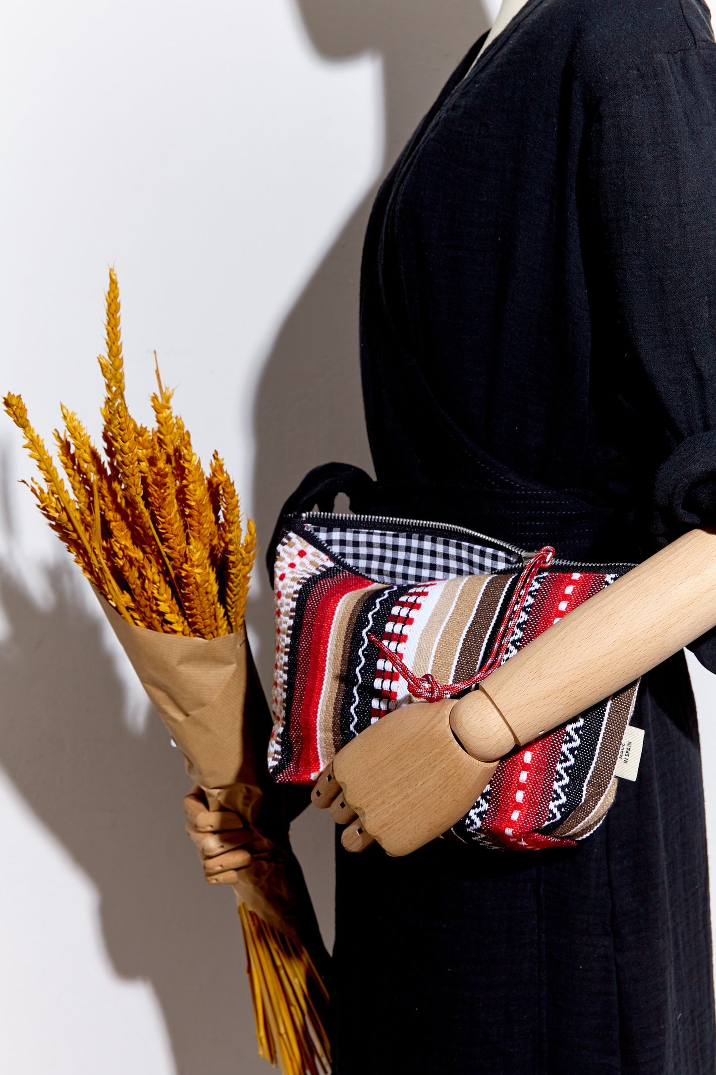 Red, black, and beige striped textile pouch with a zip closure, held by a mannequin alongside a wheat bouquet.