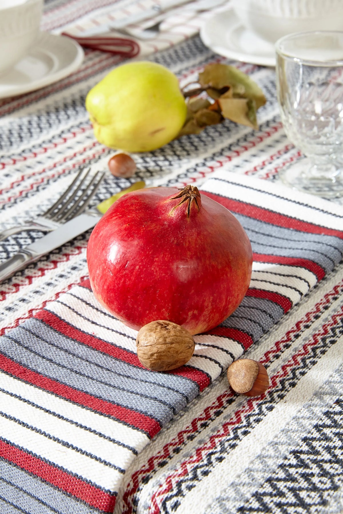Mantel de tejido tradicional con rayas granates, grises y crema sobre mesa decorada con una granada y frutos secos.