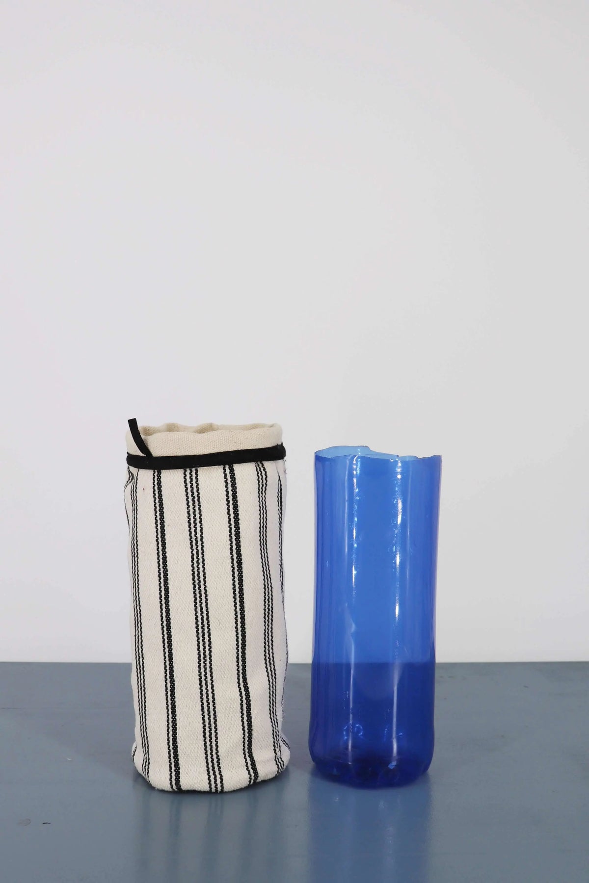 Funda de jarrón en loneta color crudo con rayas negras verticales junto a un jarrón de vidrio azul sobre fondo blanco.