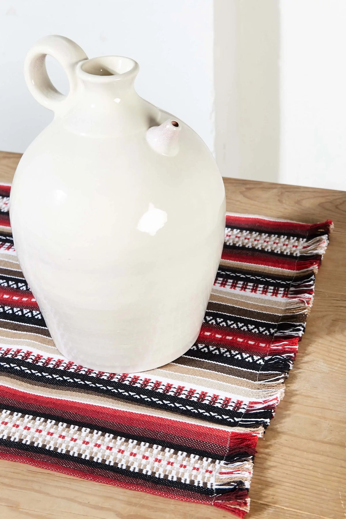 Camino de mesa tejido estilo manchego con rayas rojas, negras, beige y blancas, con flecos y un botijo blanco encima.