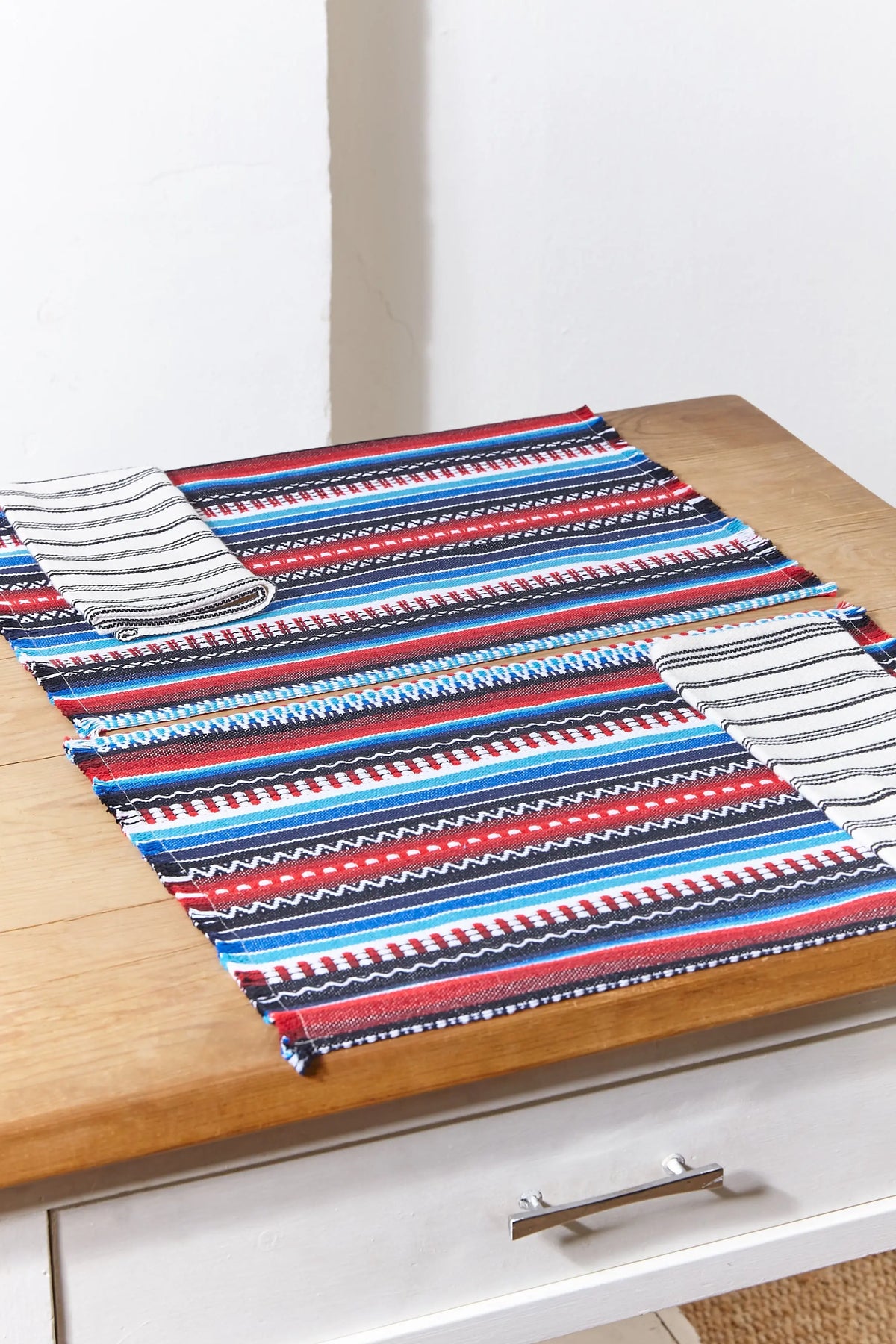 Pack de 2 manteles individuales de estilo manchego con franjas tejidas en azul, rojo y blanco sobre mesa de madera.