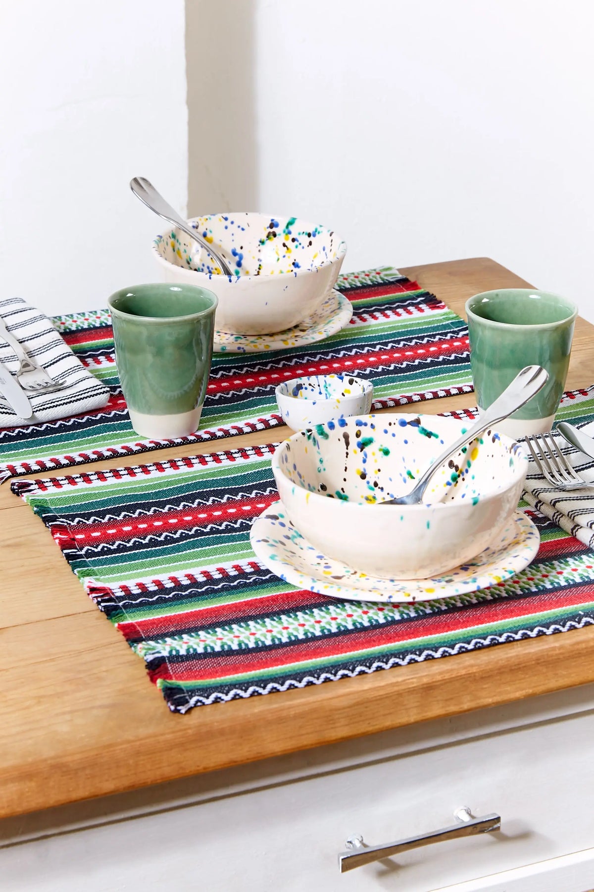 Pack of two rectangular woven placemats in green, red, and white stripes, styled on a wooden table with ceramic bowls.