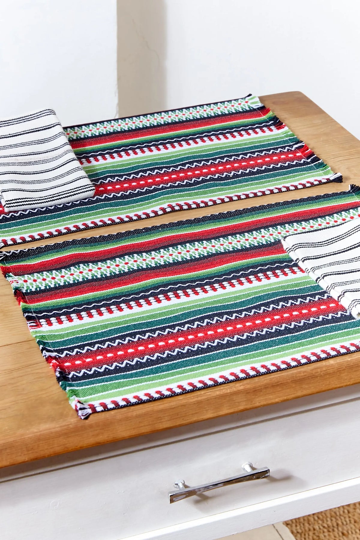 Pack de dos manteles individuales de tejido manchego con rayas horizontales en verde, rojo y blanco sobre mesa madera.