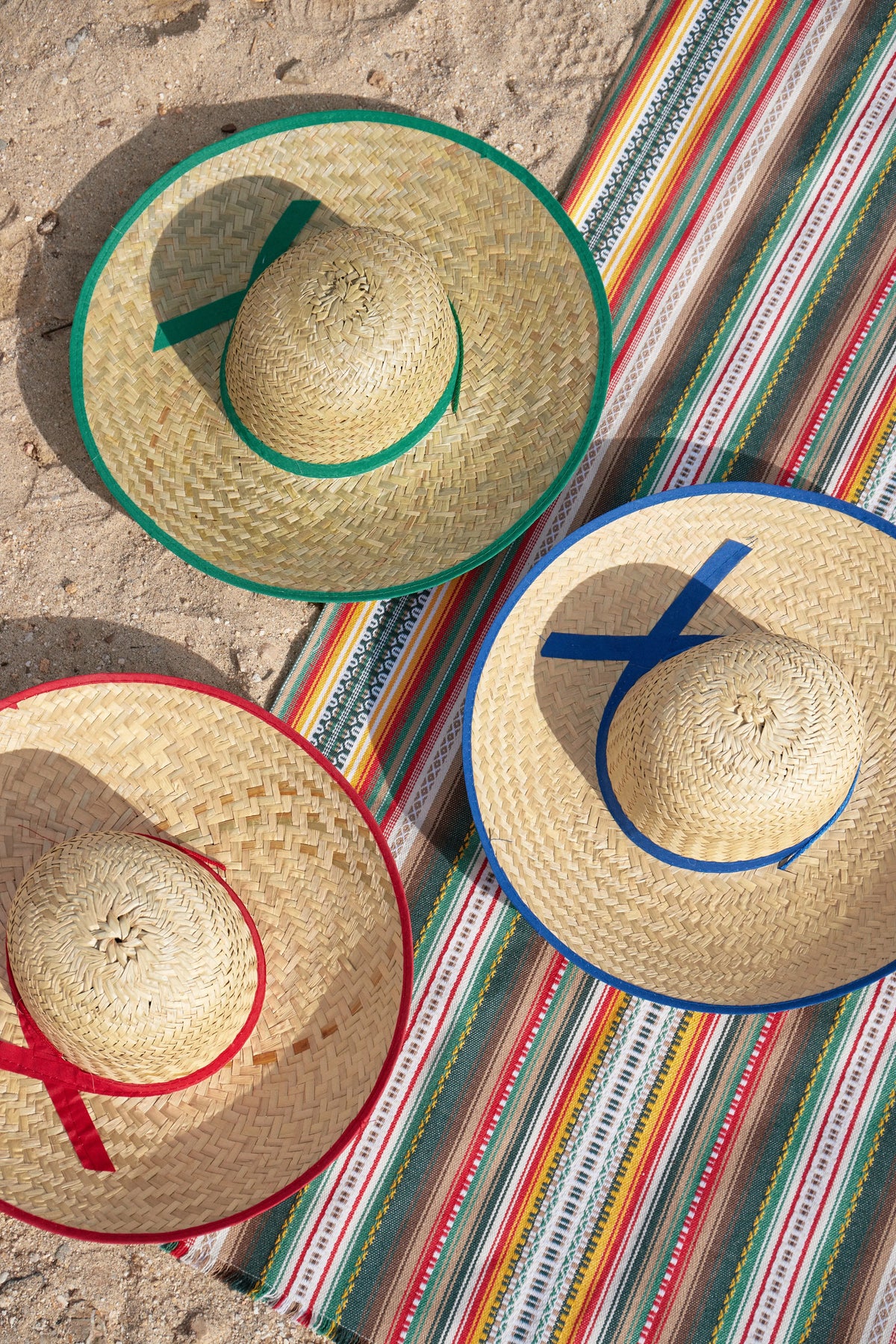 Sombreros de paja, colores verde, rojo y azul, sobre manta de rayas, ideal para días soleados y fiestas al aire libre.