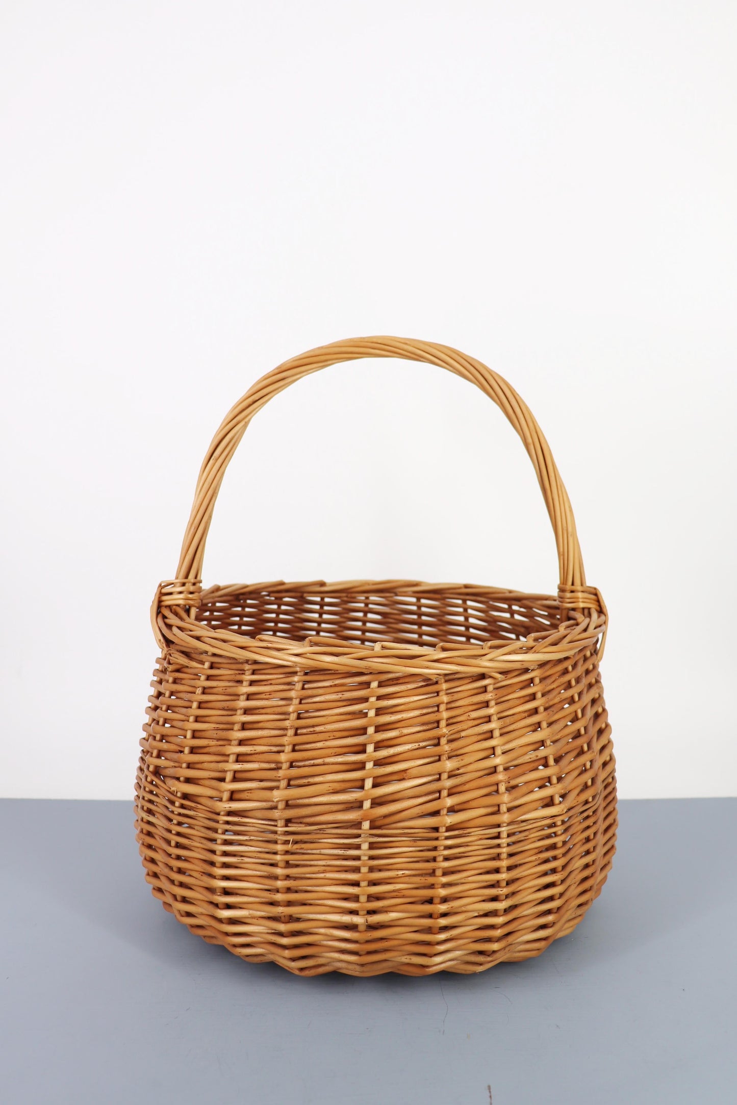 Cesta redonda de mimbre natural con trenzado horizontal y asa superior fija sobre fondo neutro.