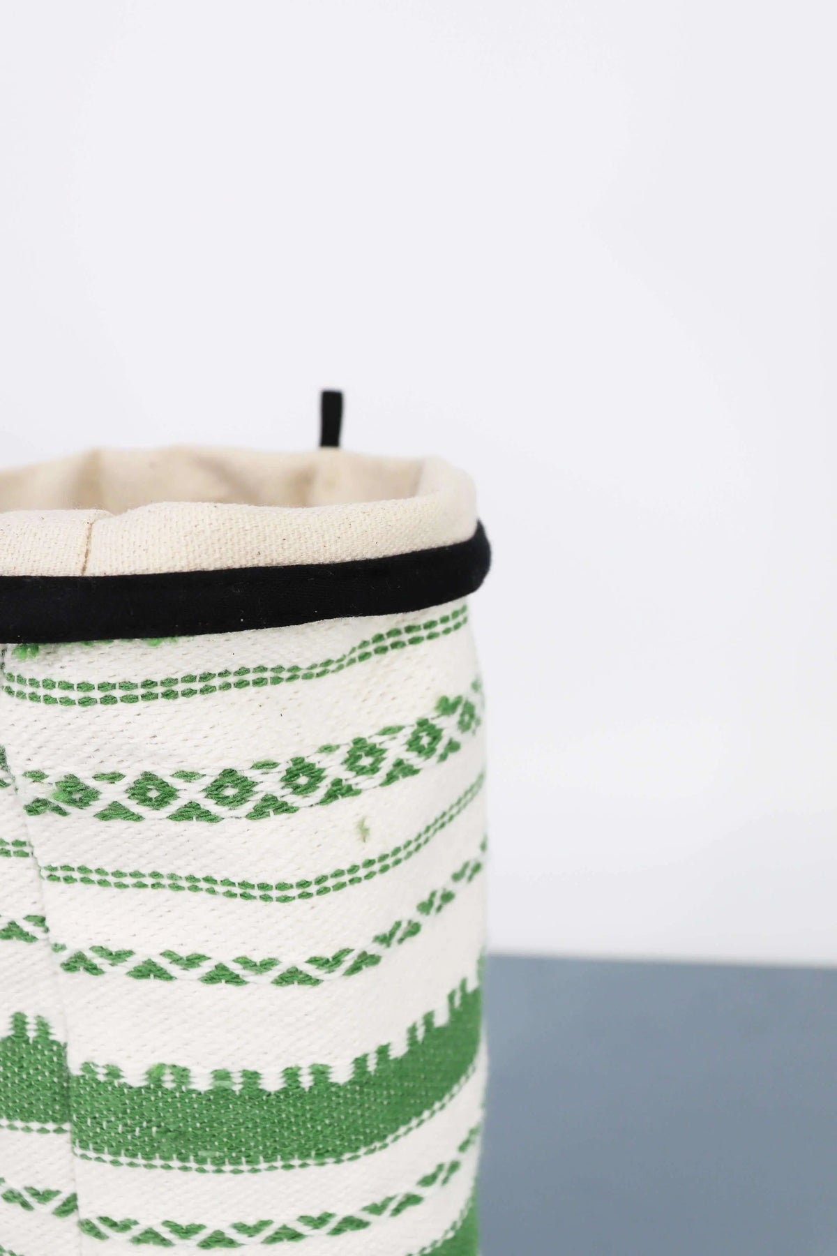 Funda para jarrón de tejido artesanal en blanco y verde con diseño geométrico y borde superior negro sobre fondo claro.
