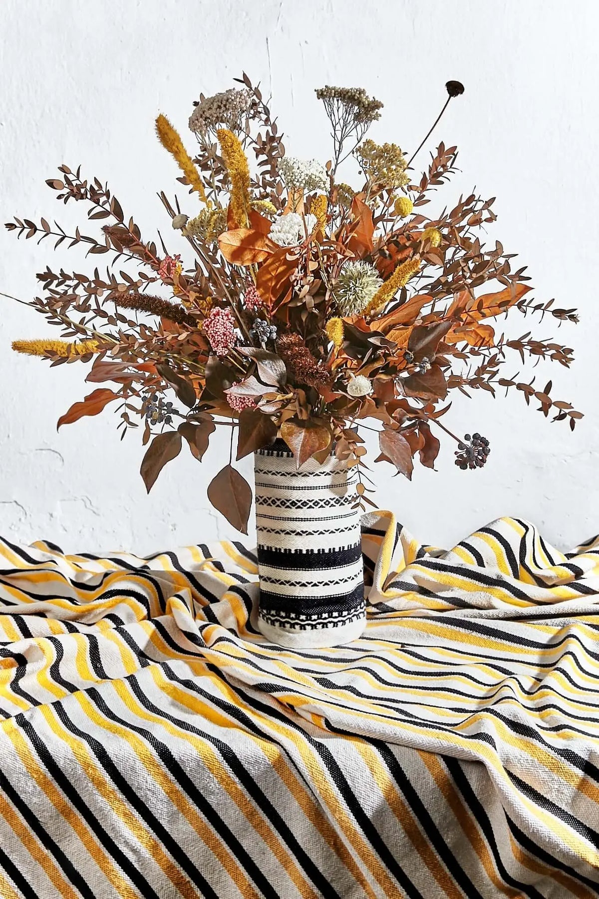 Black and white patterned textile vase cover holding a lush, autumnal arrangement of dried flowers and leaves.
