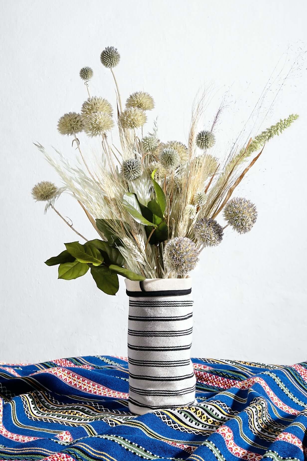 Funda de jarrón de loneta blanca con rayas negras horizontales, conteniendo un ramo de flores secas sobre tela azul.