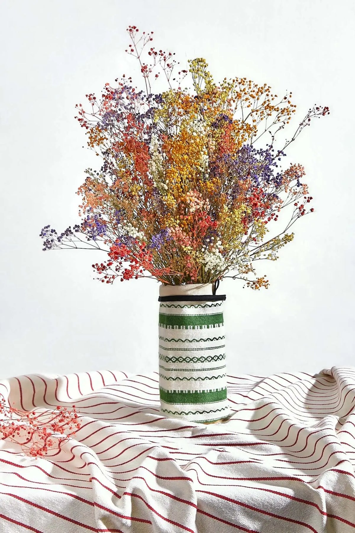 Funda para jarrón en tejido alpujarreño verde y blanco con un ramo de flores preservadas sobre mantel de rayas.