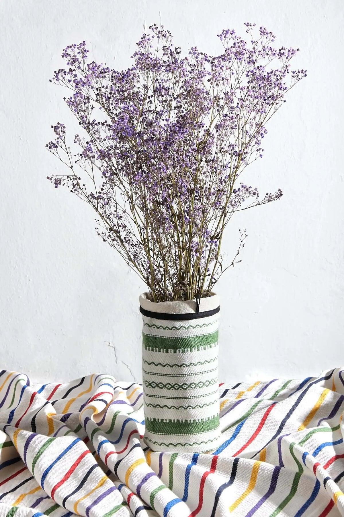 Funda para jarrón de tela blanca y verde con patrones geométricos, conteniendo un ramo de flores secas moradas.