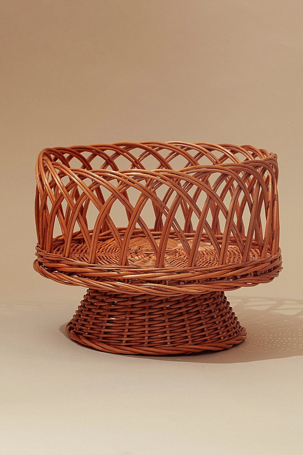 Round pedestal fruit bowl made of natural woven wicker with an openwork lattice design in a warm tan color.