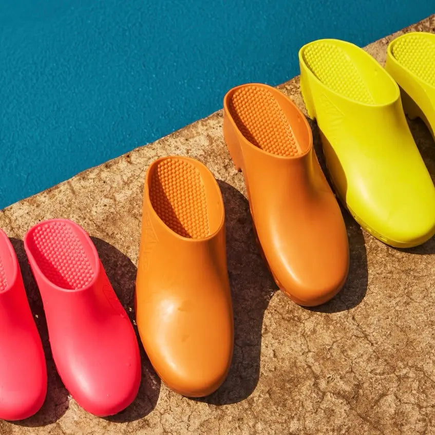 Bright orange, hole-free rubber clogs with a textured, studded insole, displayed on a stone surface by a blue pool.