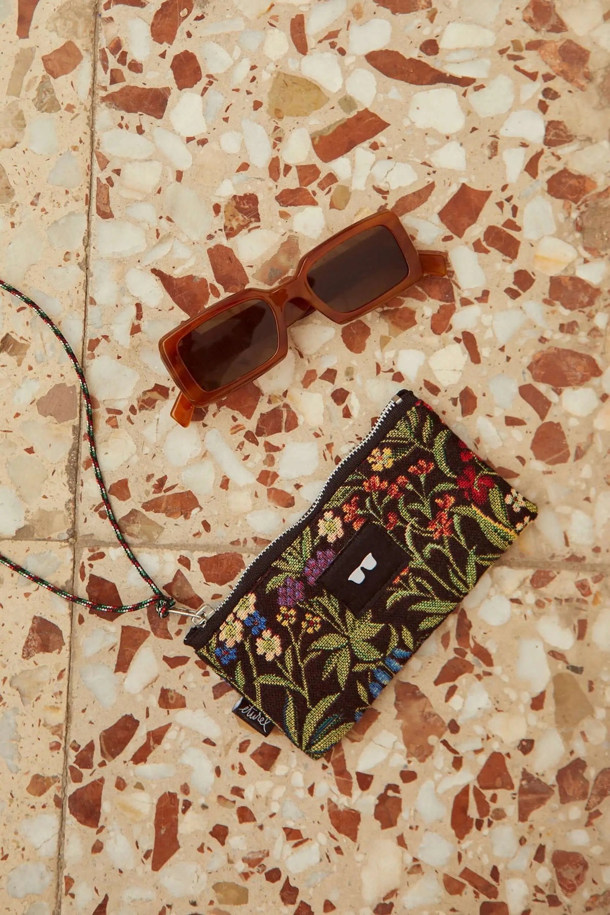 Funda para gafas de tejido jacquard con flores coloridas y cordón, junto a gafas de sol sobre suelo de terrazo.