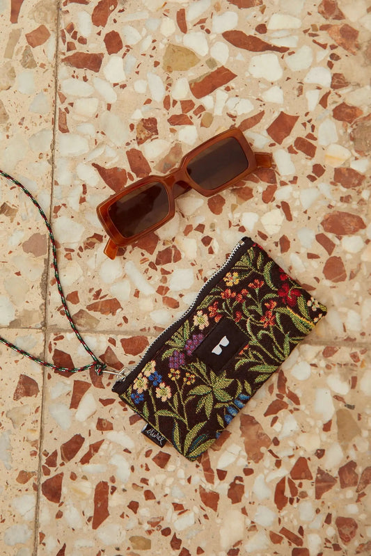 Funda para gafas de tejido jacquard con flores coloridas y cordón, junto a gafas de sol sobre suelo de terrazo.