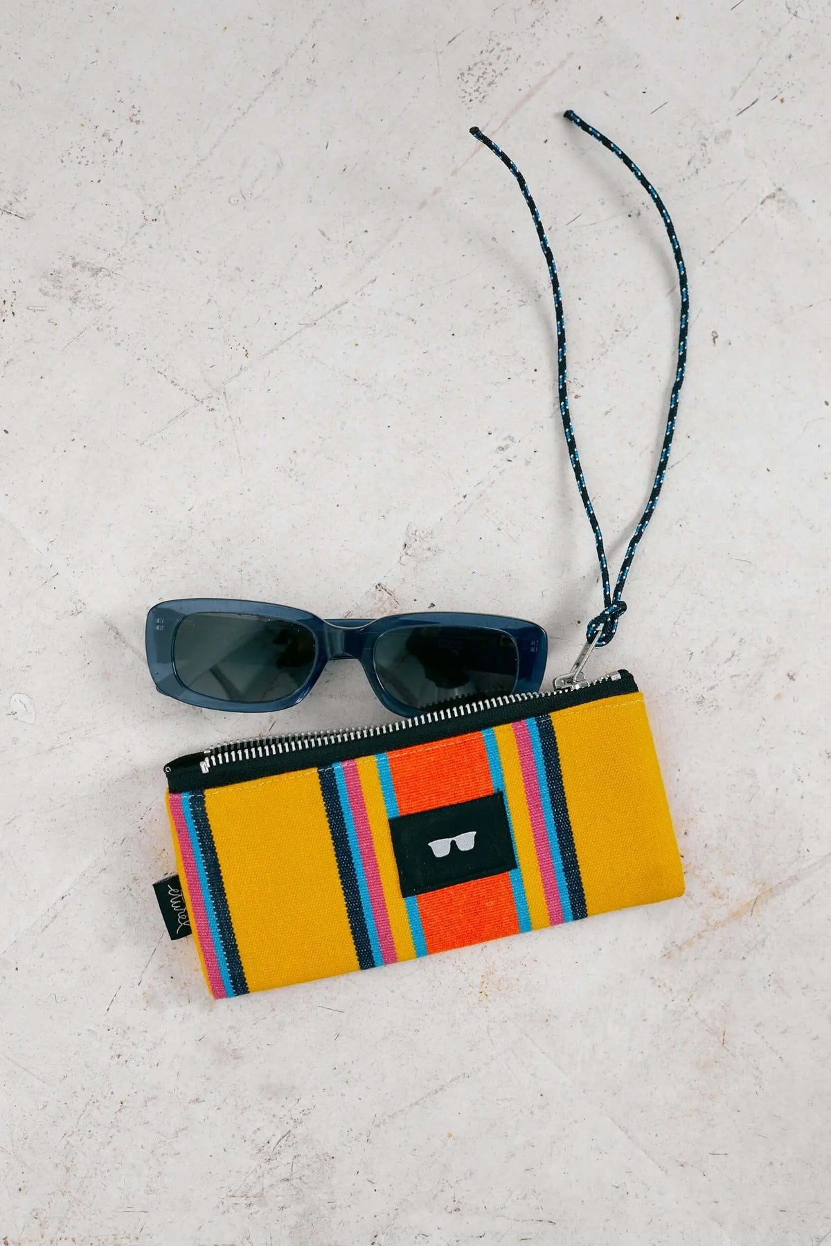 Funda para gafas de lona tipo hamaca en color amarillo a rayas con cierre de cremallera y cordón, junto a unas gafas.