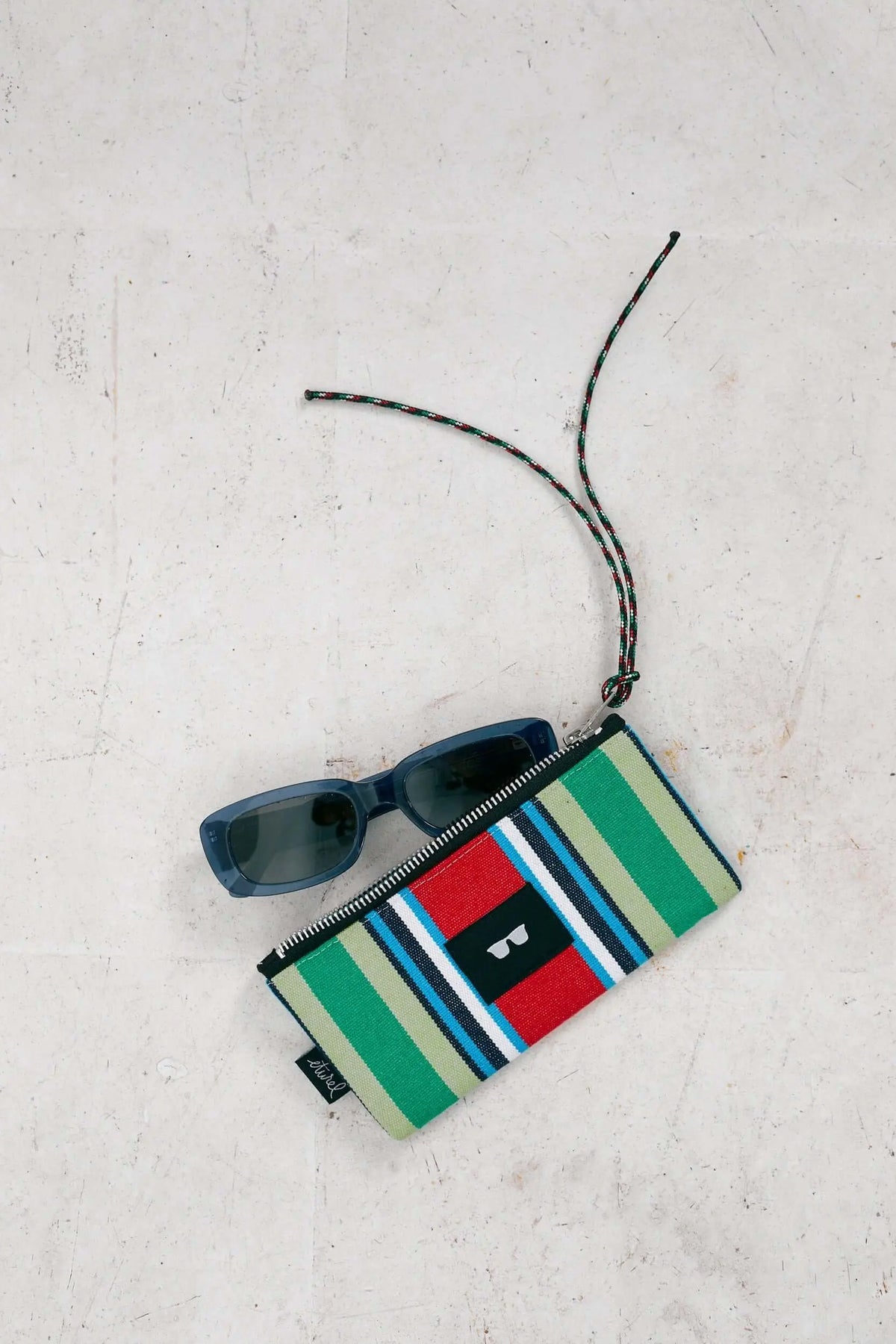 Funda de gafas de lona a rayas rojas y verdes con cordón y cremallera, junto a unas gafas de sol oscuras.