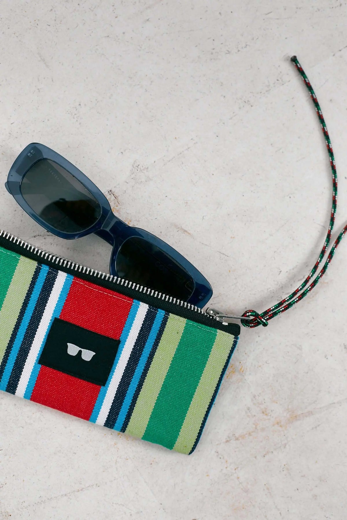 Funda de lona a rayas rojas, verdes y azules con cierre de cremallera y cordón, junto a gafas de sol azules.
