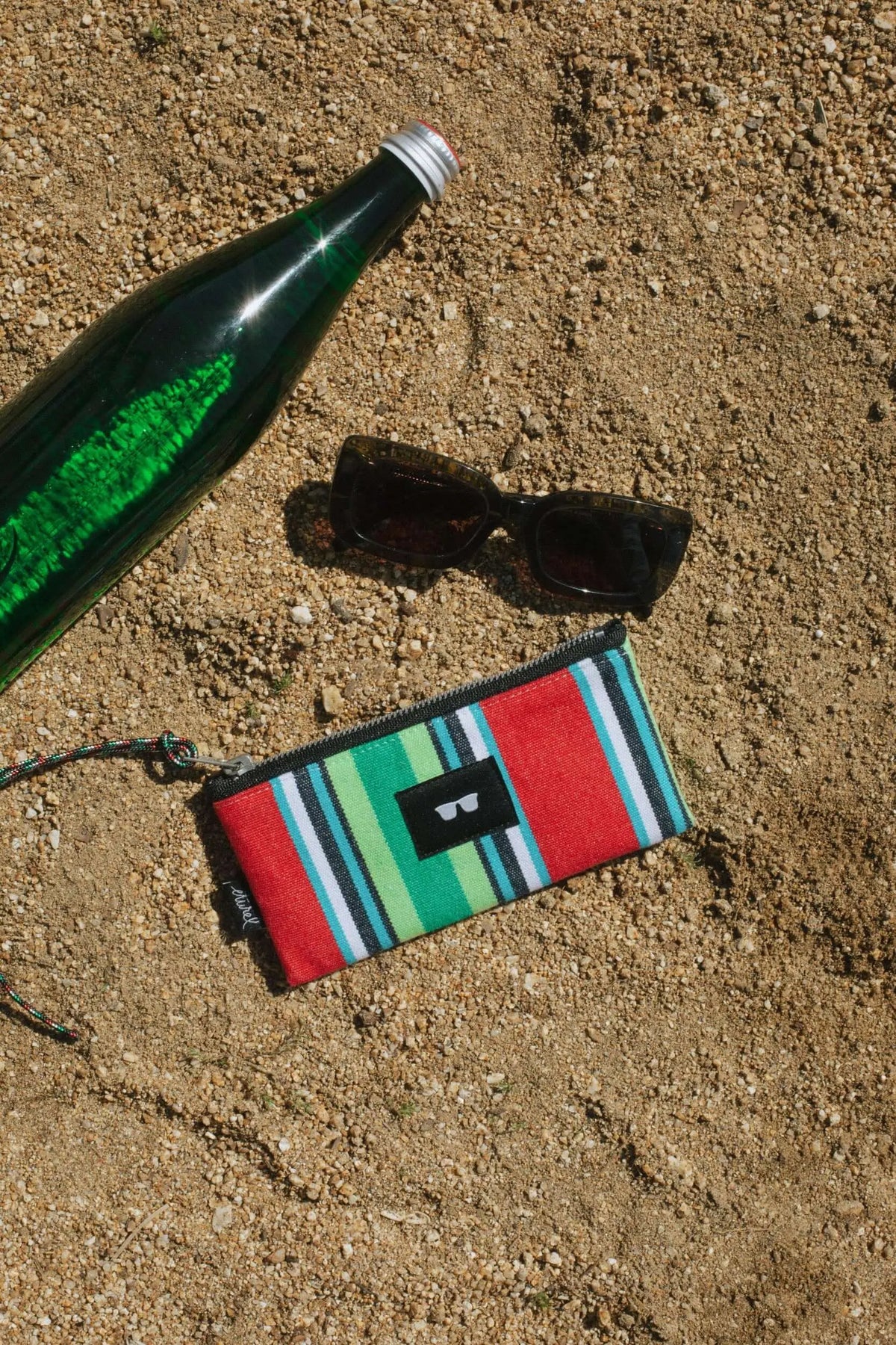 Funda de lona a rayas rojas y verdes con cremallera, junto a unas gafas de sol y botella sobre arena.