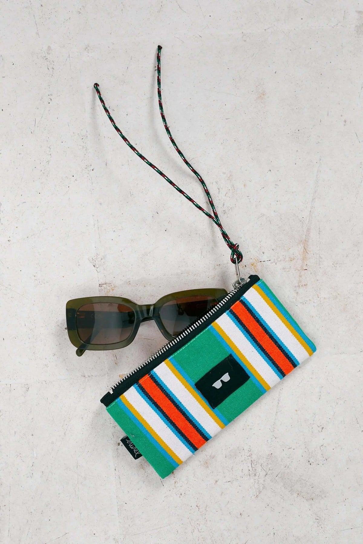 Funda de gafas de lona a rayas verdes, naranjas y blancas con cremallera y cordón, junto a unas gafas de sol verdes.