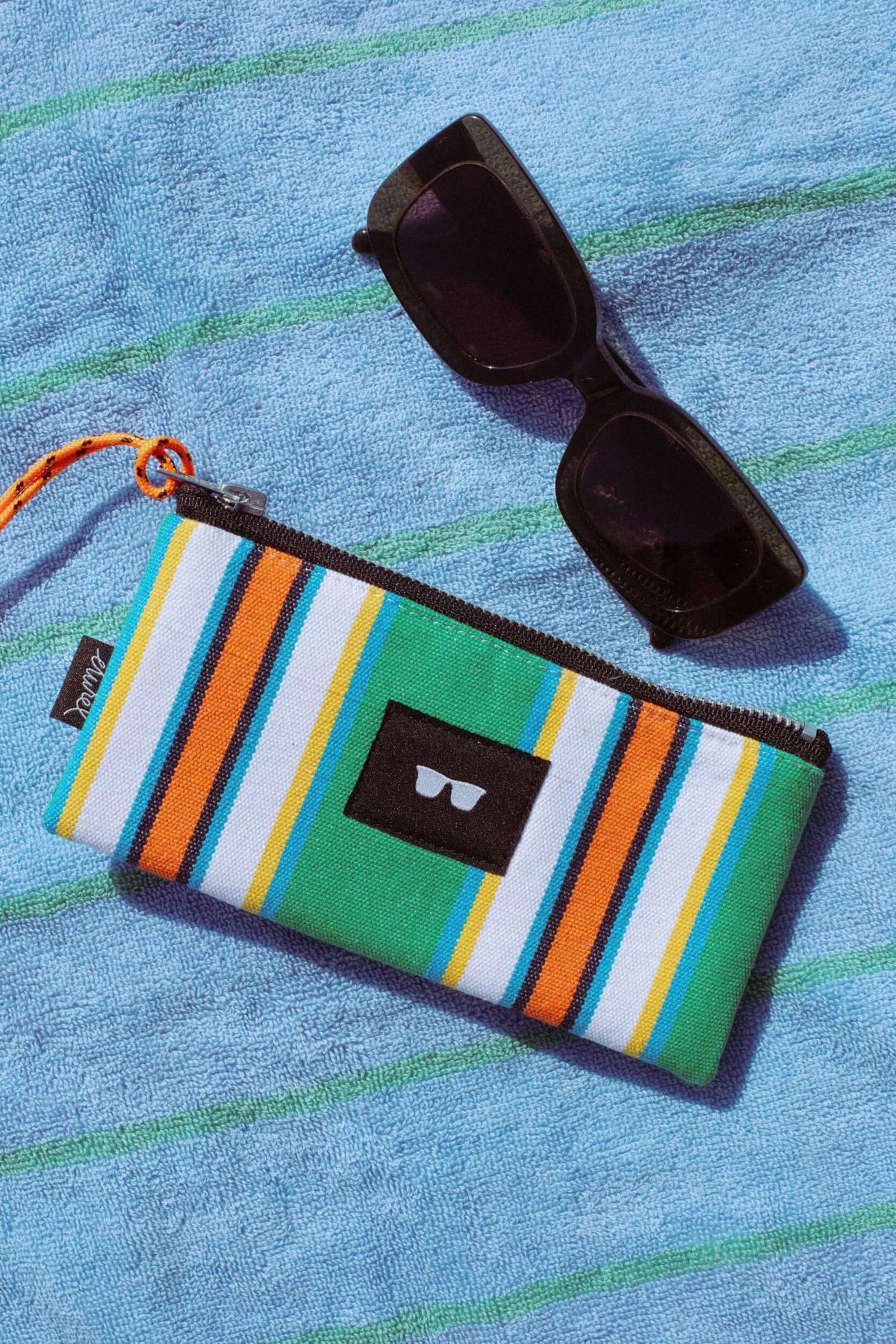 Funda de lona a rayas verdes, naranjas y blancas con cremallera, junto a gafas de sol sobre una toalla azul.