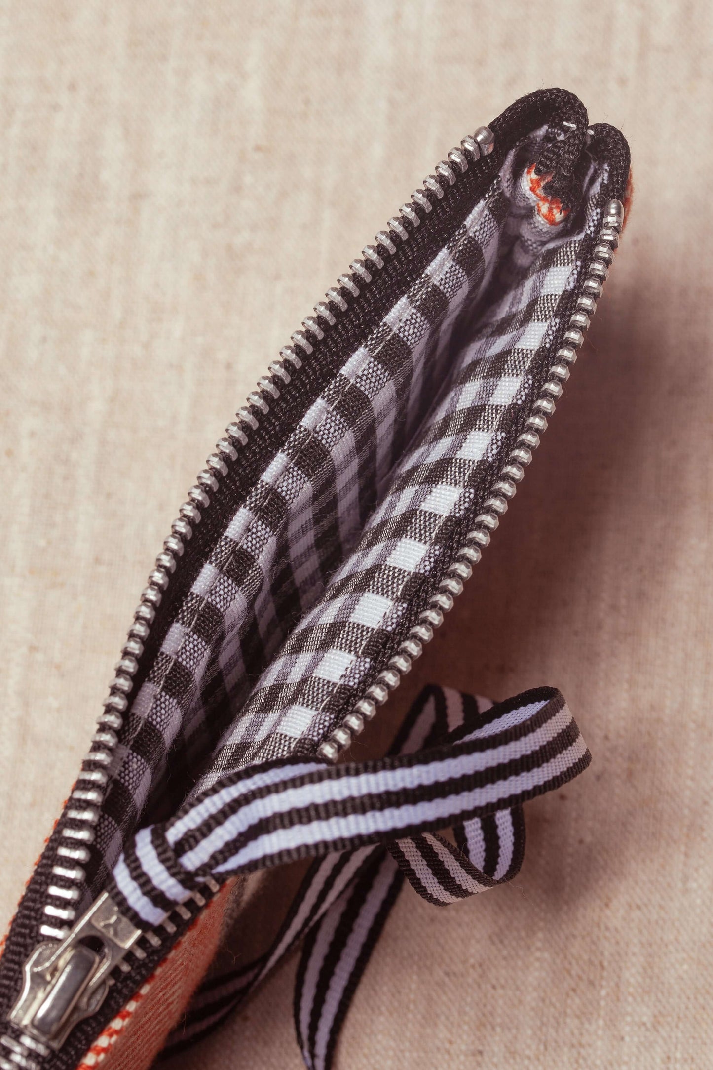 Clay-colored twill glasses case with a zipper, black and white gingham lining, and a striped ribbon pull.