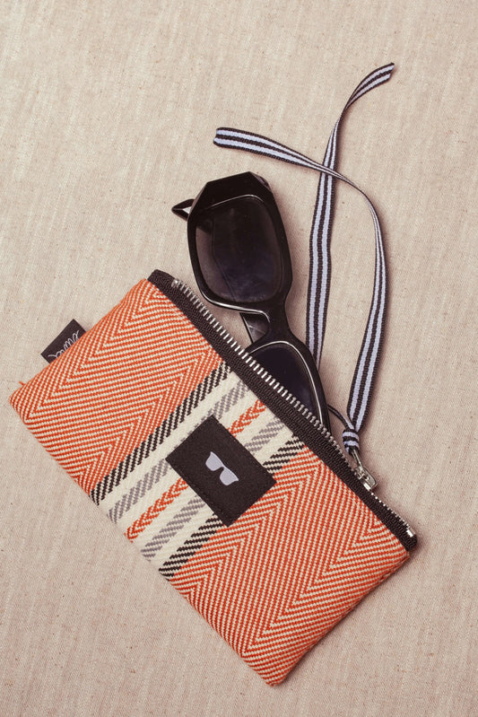Clay-colored herringbone glasses case with a zipper, black accent label, and striped strap holding black sunglasses.
