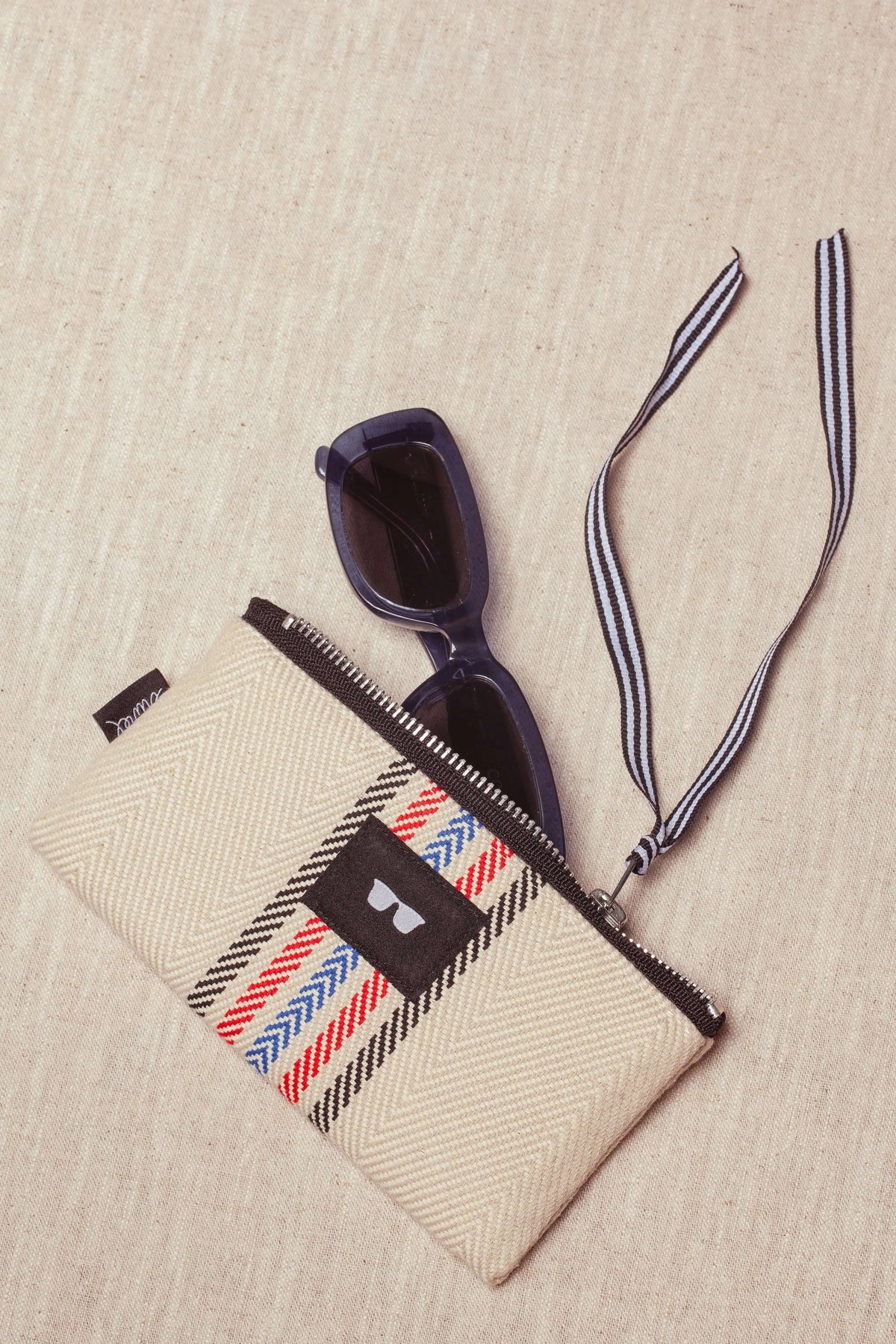 Funda de gafas de sarga color crudo con patrón de espiga, cremallera, cinta rayada y gafas de sol oscuras dentro.
