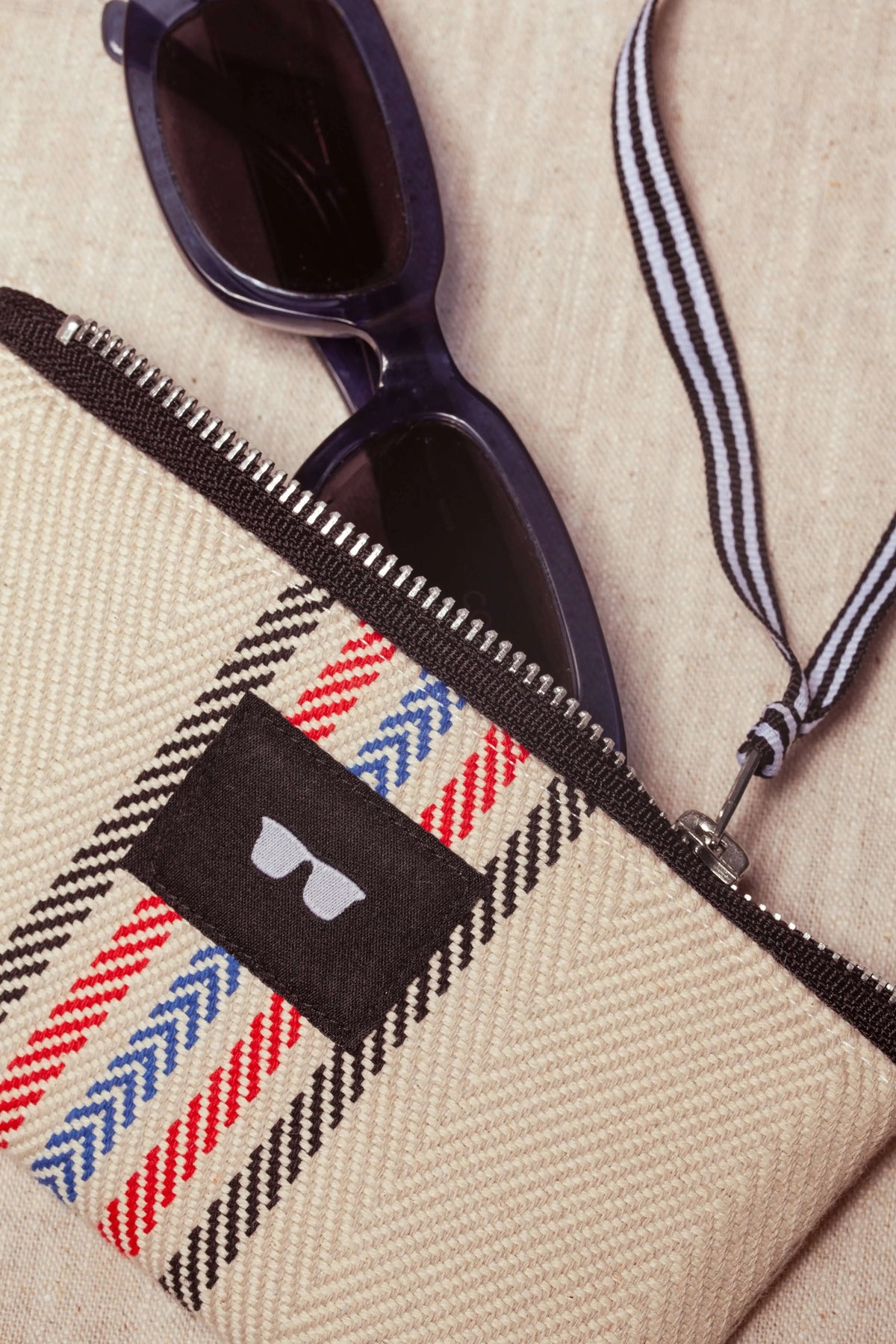 Funda de tela cruda con patrón de espiga, rayas rojas y azules, cierre de cremallera y cordón, conteniendo gafas oscuras.