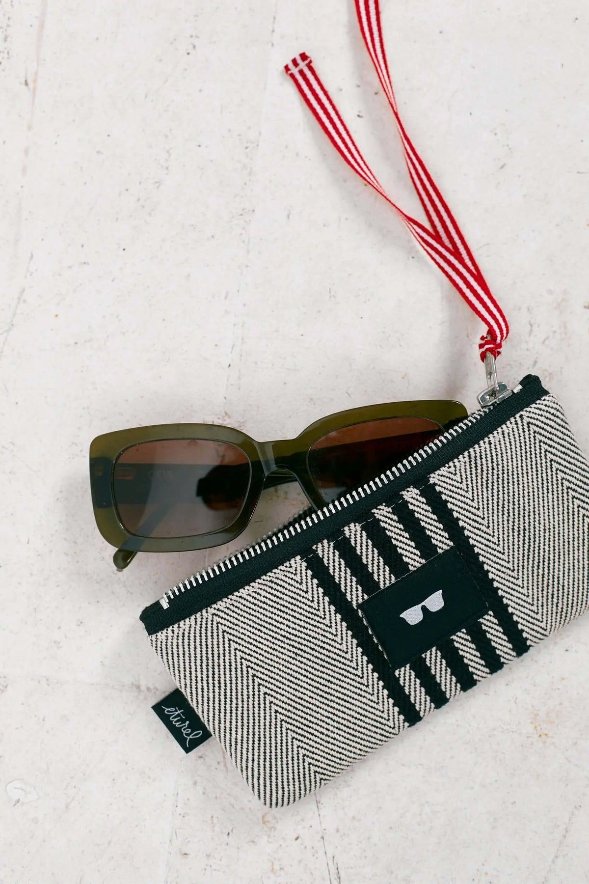 Funda de tela sarga negra con patrón en espiga blanco, cremallera, cinta roja a rayas y gafas de sol verde oliva.