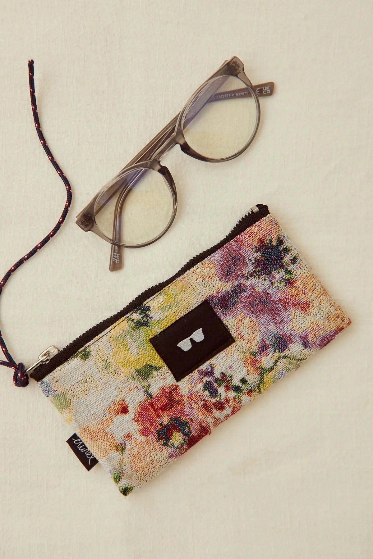 Glasses case in floral jacquard fabric with a zip, brown accent patch, and attached cord next to a pair of glasses.