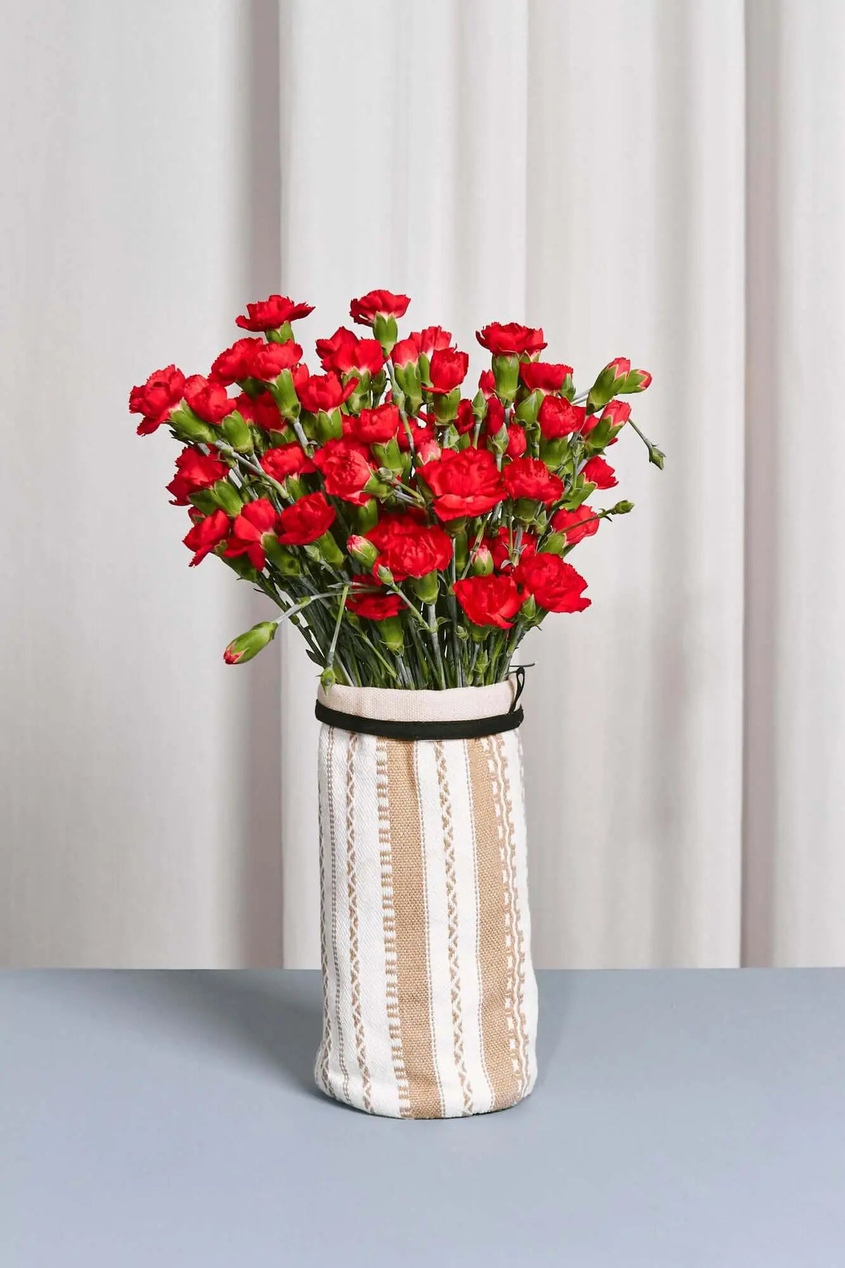 Funda de jarrón de tela cruda y blanca con rayas verticales, conteniendo un ramo de claveles rojos brillantes.