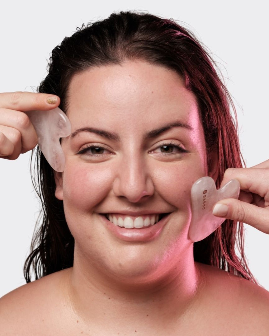 Mujer sonriente usando dos piedras Gua Sha de cuarzo rosa suave y translúcido de la marca Banbu sobre su rostro.