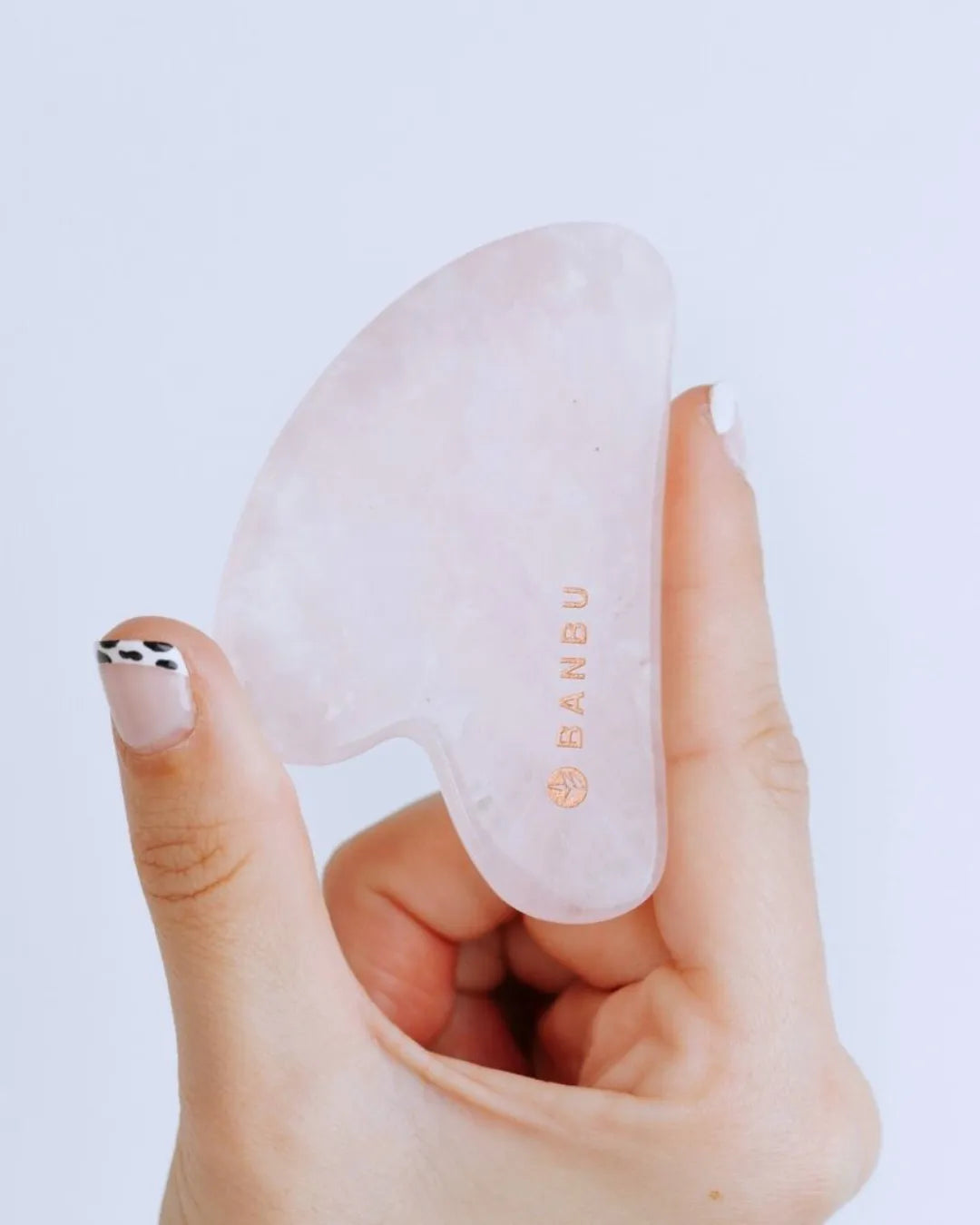 Mano sosteniendo un Gua Sha de cuarzo rosa natural, translúcido y suave, con el logo de la marca BANBU grabado.