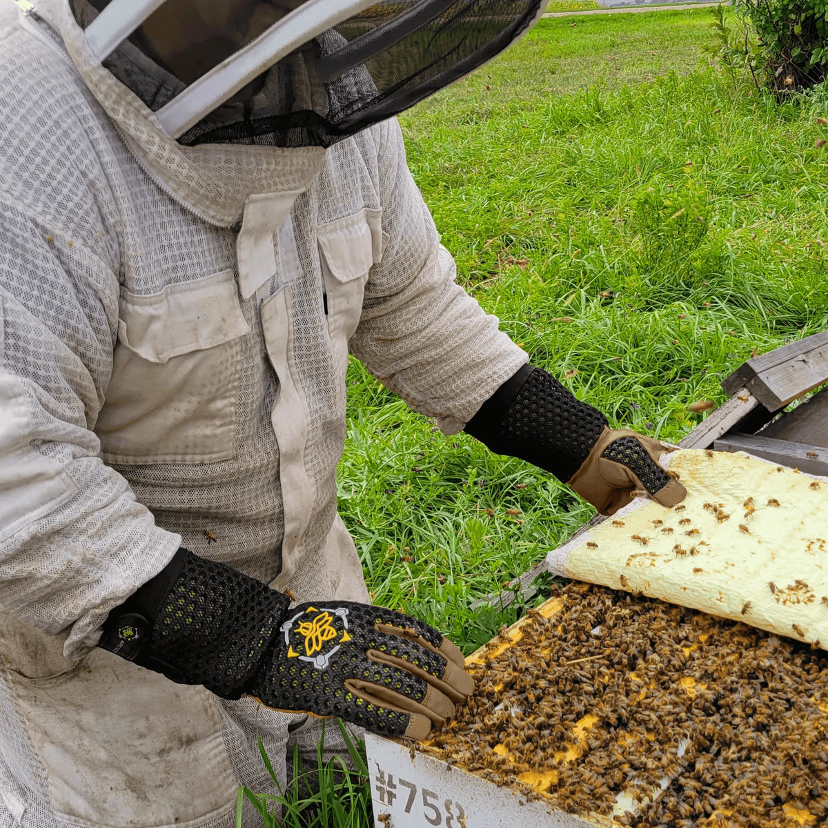 Beekeeper wearing black, heavy-duty tactical gloves with tan leather palms and ventilated mesh backs over a white suit.