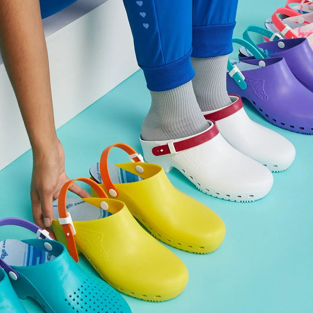 Yellow rubber clog without holes, featuring an orange pivoting heel strap, set against a colorful row of similar shoes.