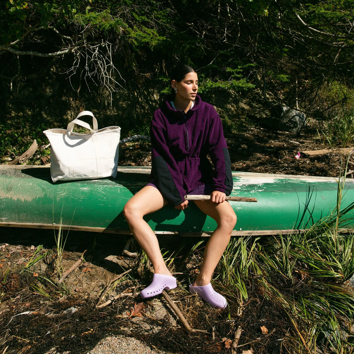 Lavender, rubber-style clogs with side ventilation holes worn by a person sitting outdoors on a green canoe.