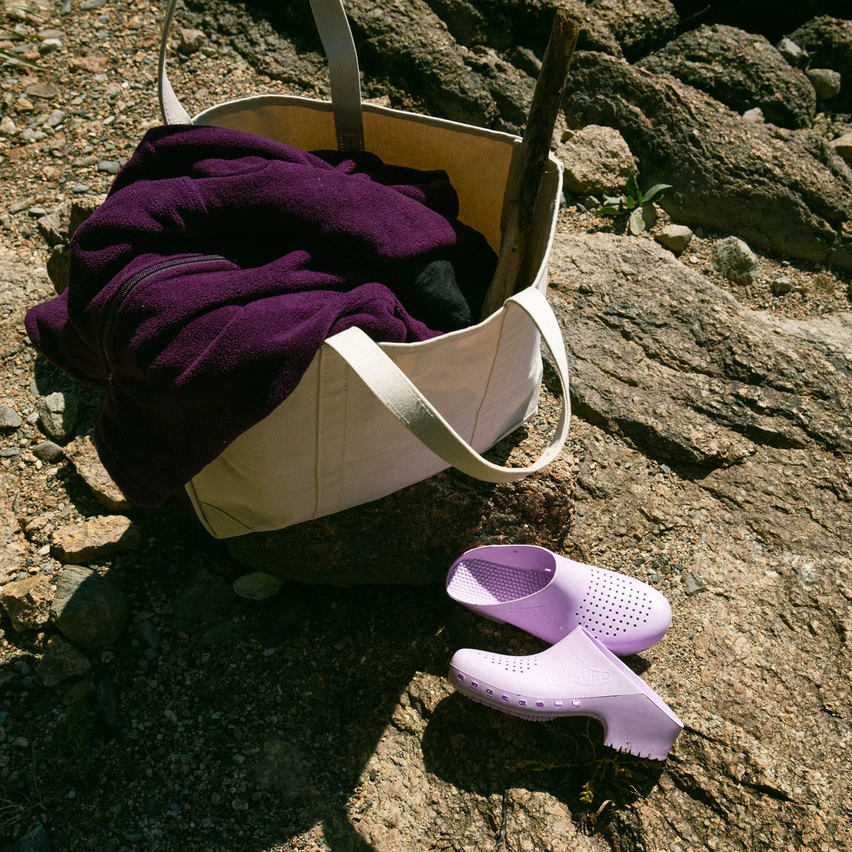 Lavender slip-on clogs with ventilation holes, set on a rocky ground next to a canvas tote bag.