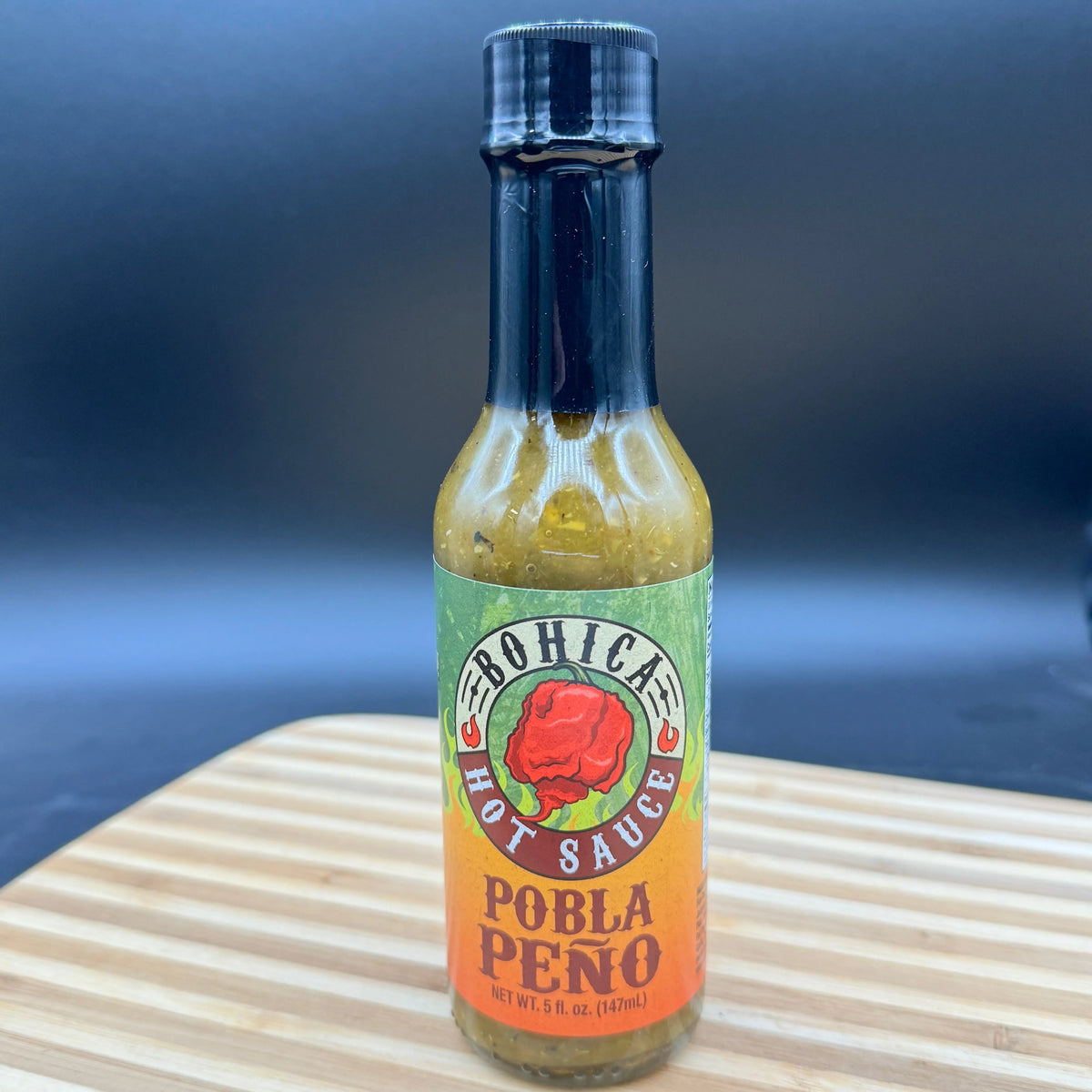 A glass bottle of green Pobla Peño hot sauce with a label featuring a red pepper, set on a wooden cutting board.
