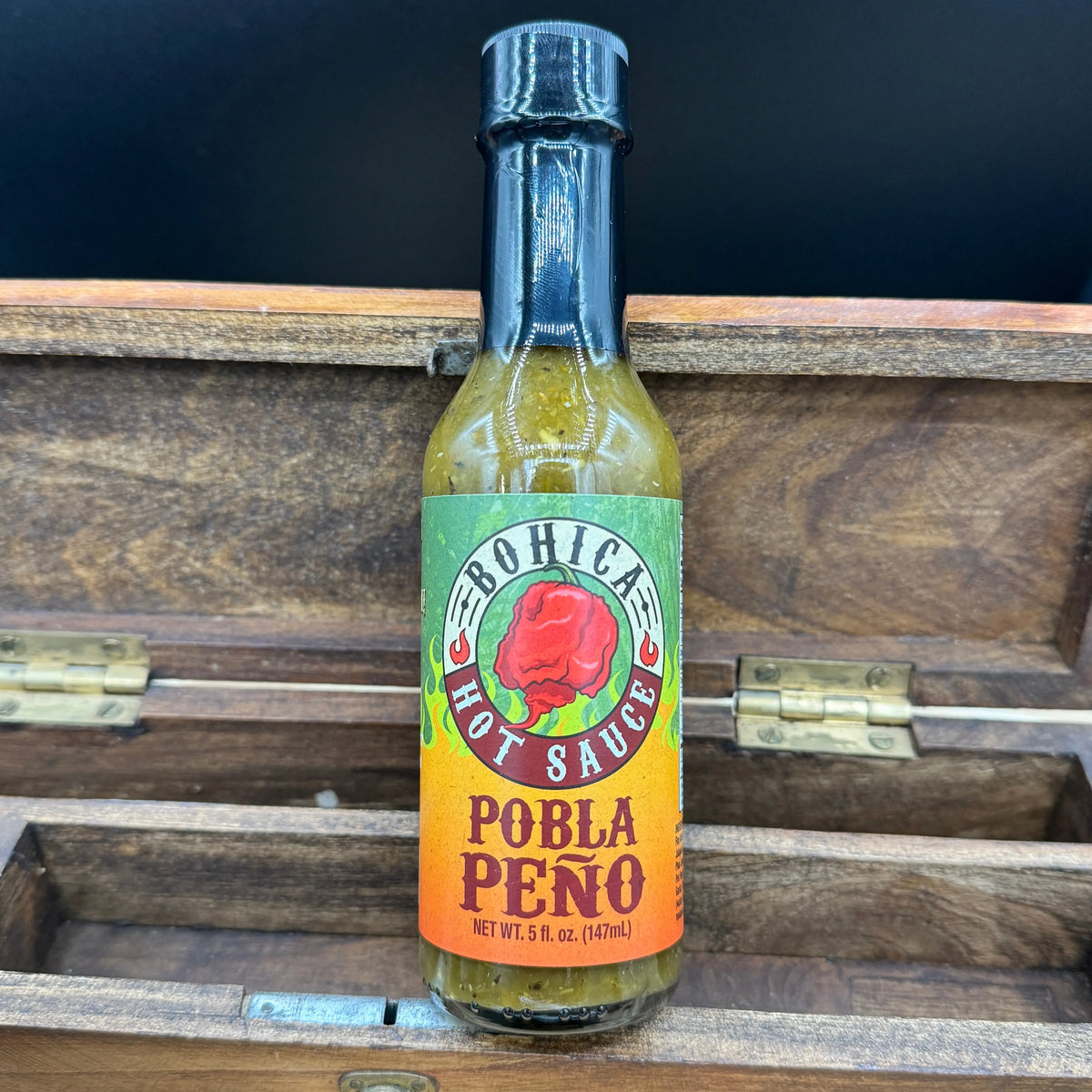 A bottle of green Pobla Peño hot sauce with a red pepper graphic on the label, standing in a wooden box.