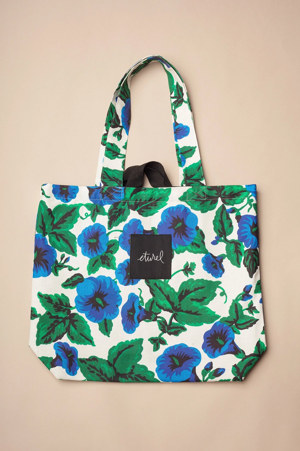 Bolso tote de lona con estampado de flores azules y hojas verdes sobre fondo blanco, asas reforzadas y estilo casual.