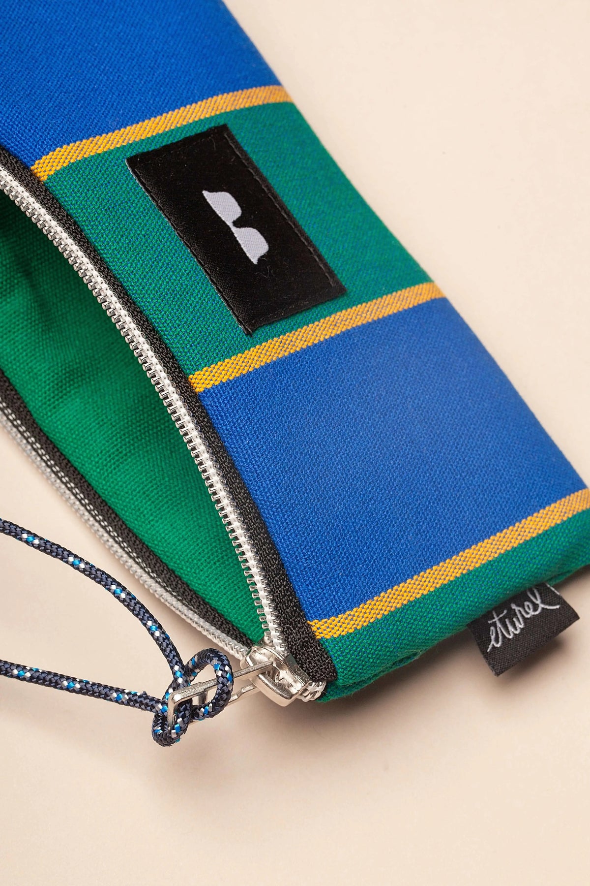 Funda de gafas en lona estilo toldo a rayas azules, verdes y amarillas con cremallera y cordón de sujeción.
