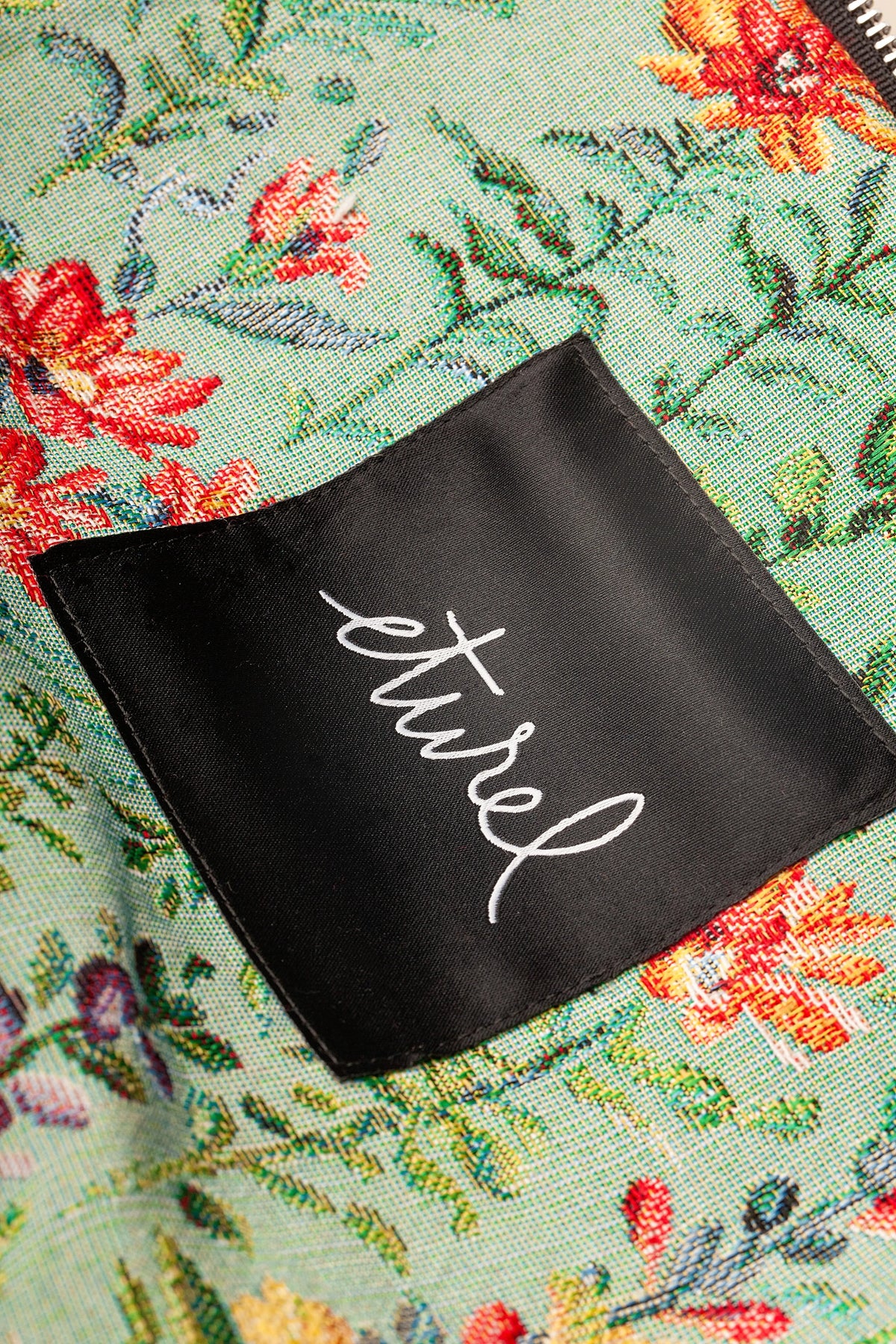 Bolso bandolera de tejido jacquard celeste con estampado floral de azaleas y etiqueta negra frontal con letras blancas.