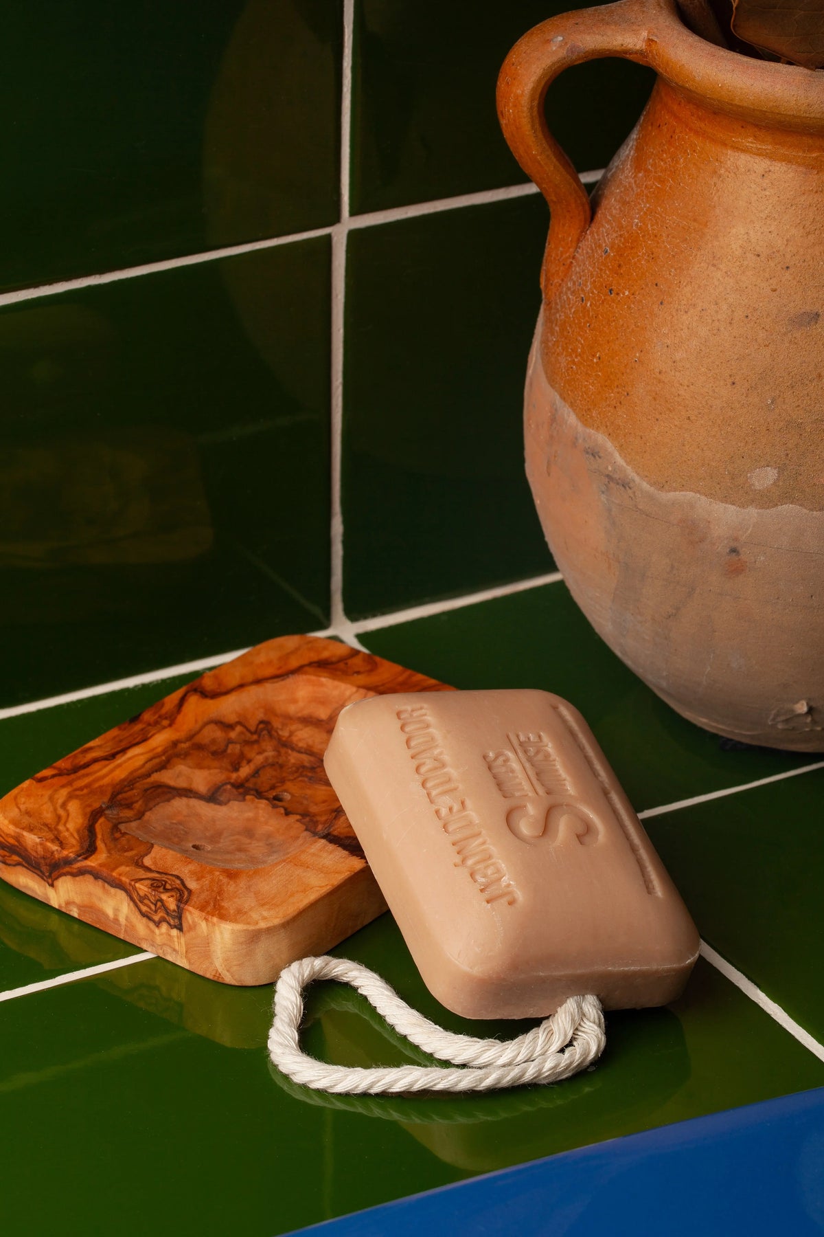 Jabonera artesanal de madera de olivo con veteado natural sobre azulejos verdes, junto a pastilla de jabón con cuerda.