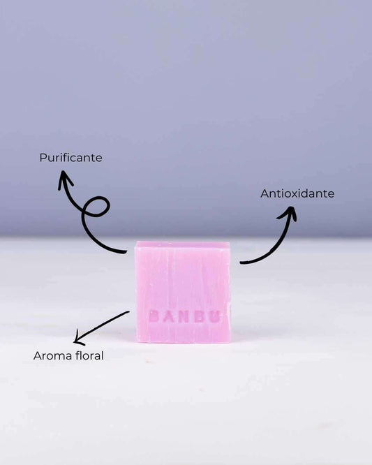 Jabón sólido rosa de Banbu con textura natural sobre fondo lila. Indica propiedades purificantes, antioxidantes y aroma.