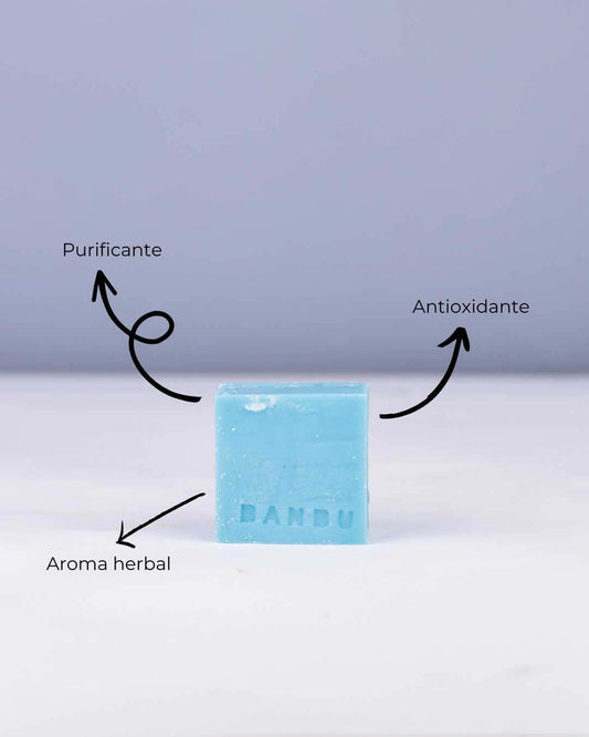 Jabón sólido azul Banbu sobre fondo claro, con flechas que señalan sus propiedades: purificante, antioxidante y herbal.
