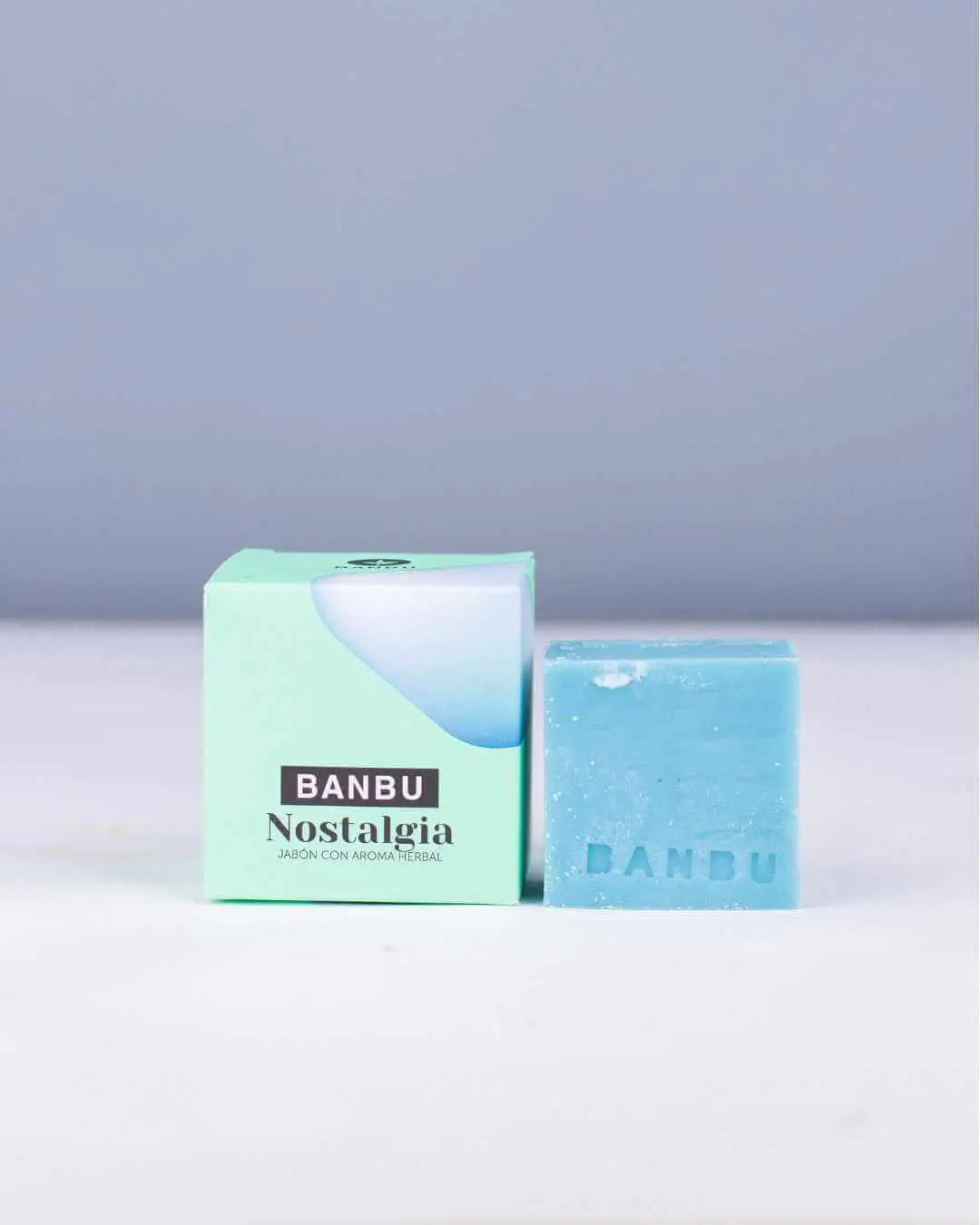 Pastilla de jabón sólido azul Banbu junto a su caja verde claro sobre fondo gris. Cosmética ecológica con aroma herbal.