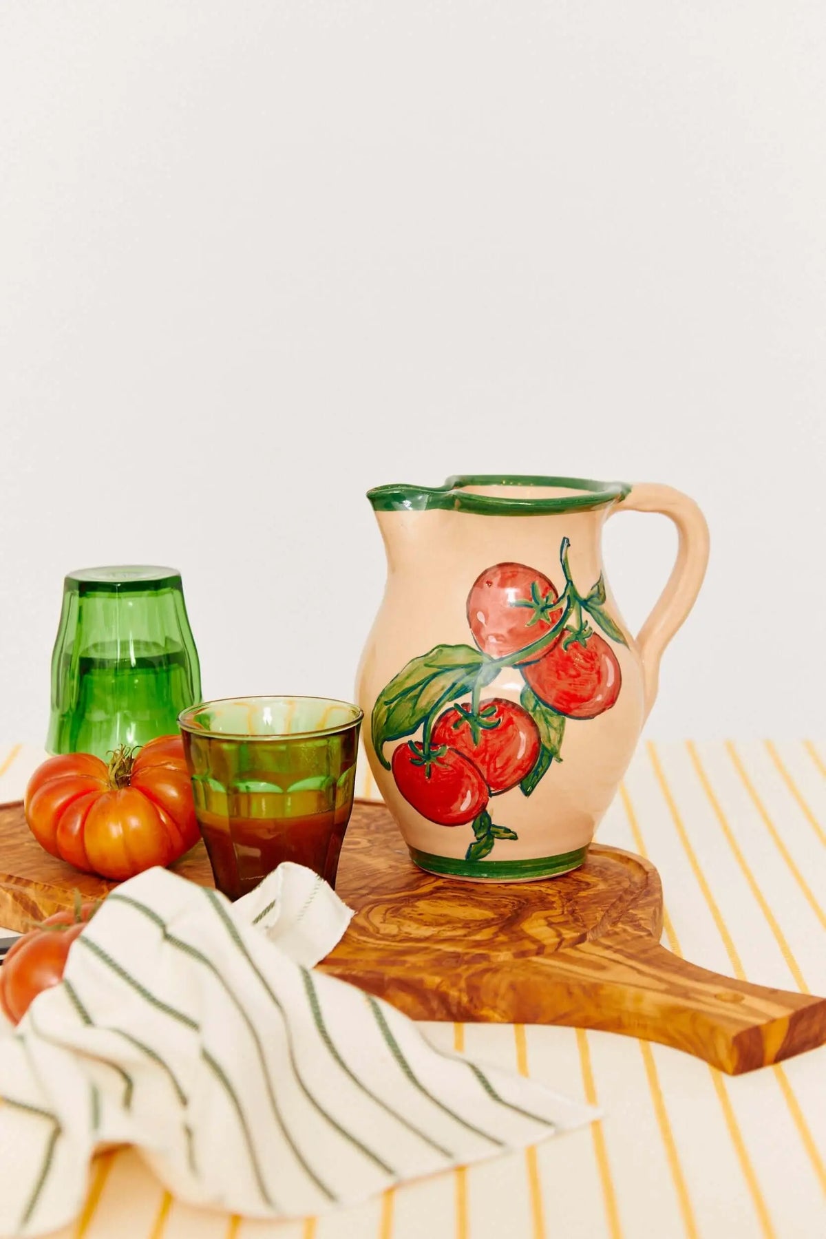 Hand-painted cream ceramic pitcher with a red tomato motif, green rim, and handle on a wooden board.